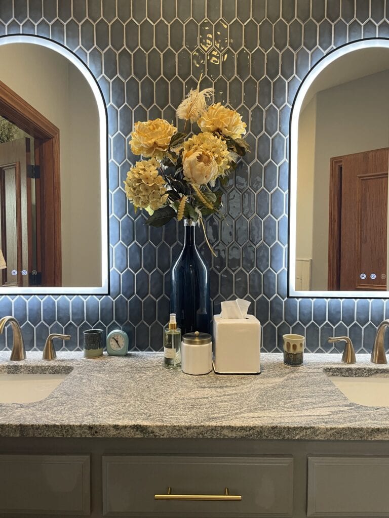 Bathroom remodel with gray vanity, stone countertop, lighted mirrors, tile backsplash, and brushed gold fixtures.